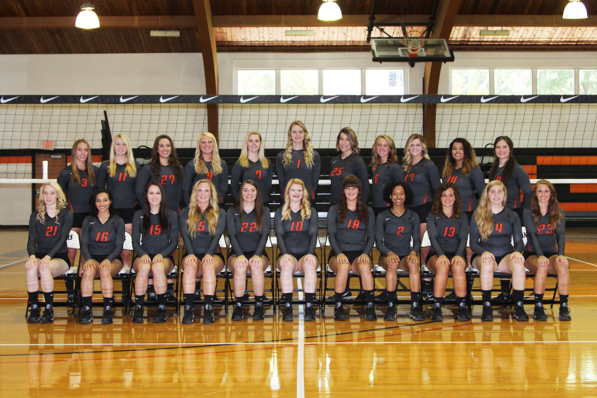 Greenville women's volleyball team photo in gray and orange kits in front of the net.