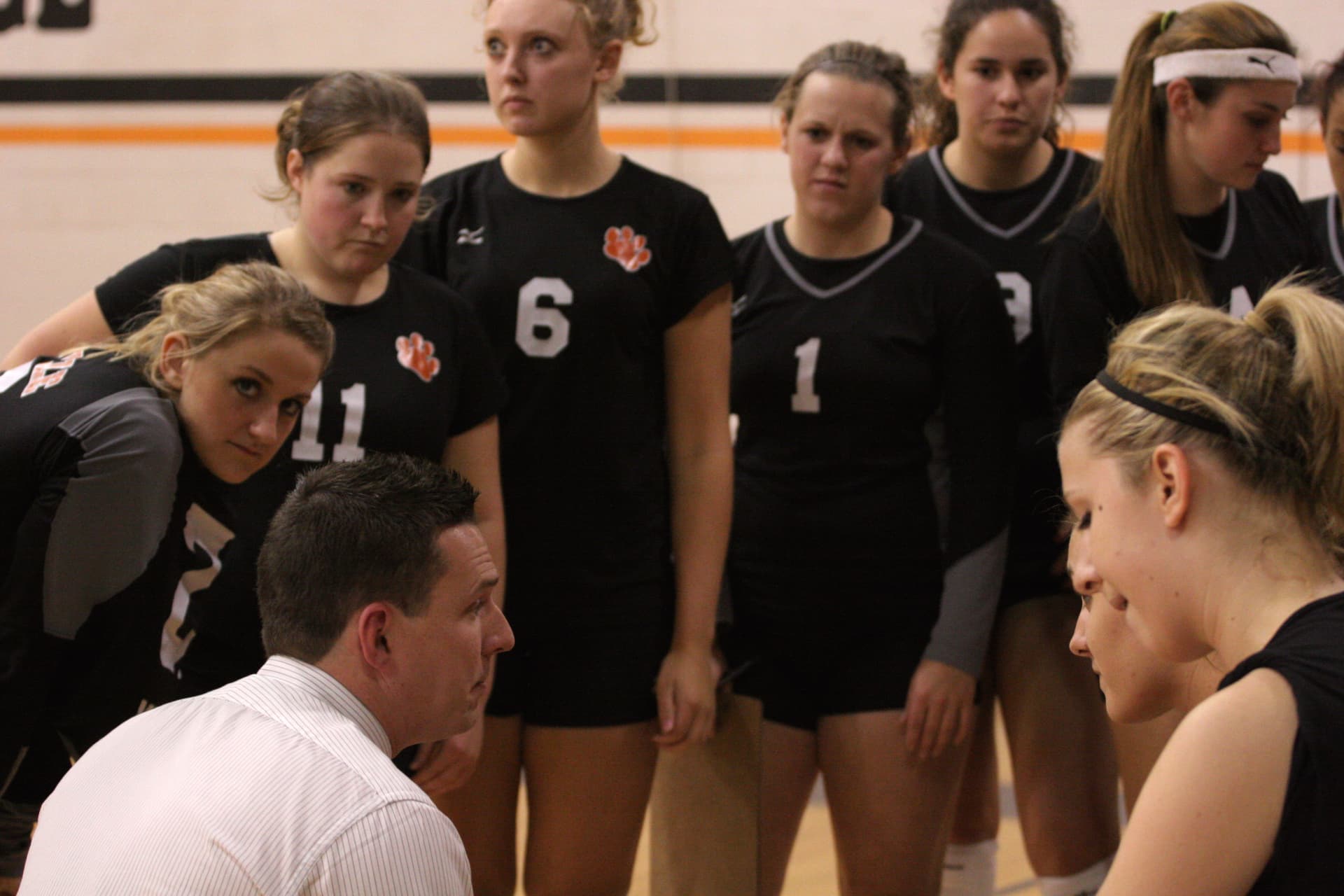 Greenville women's volleyball players in black jerseys listening to their coach courtside.