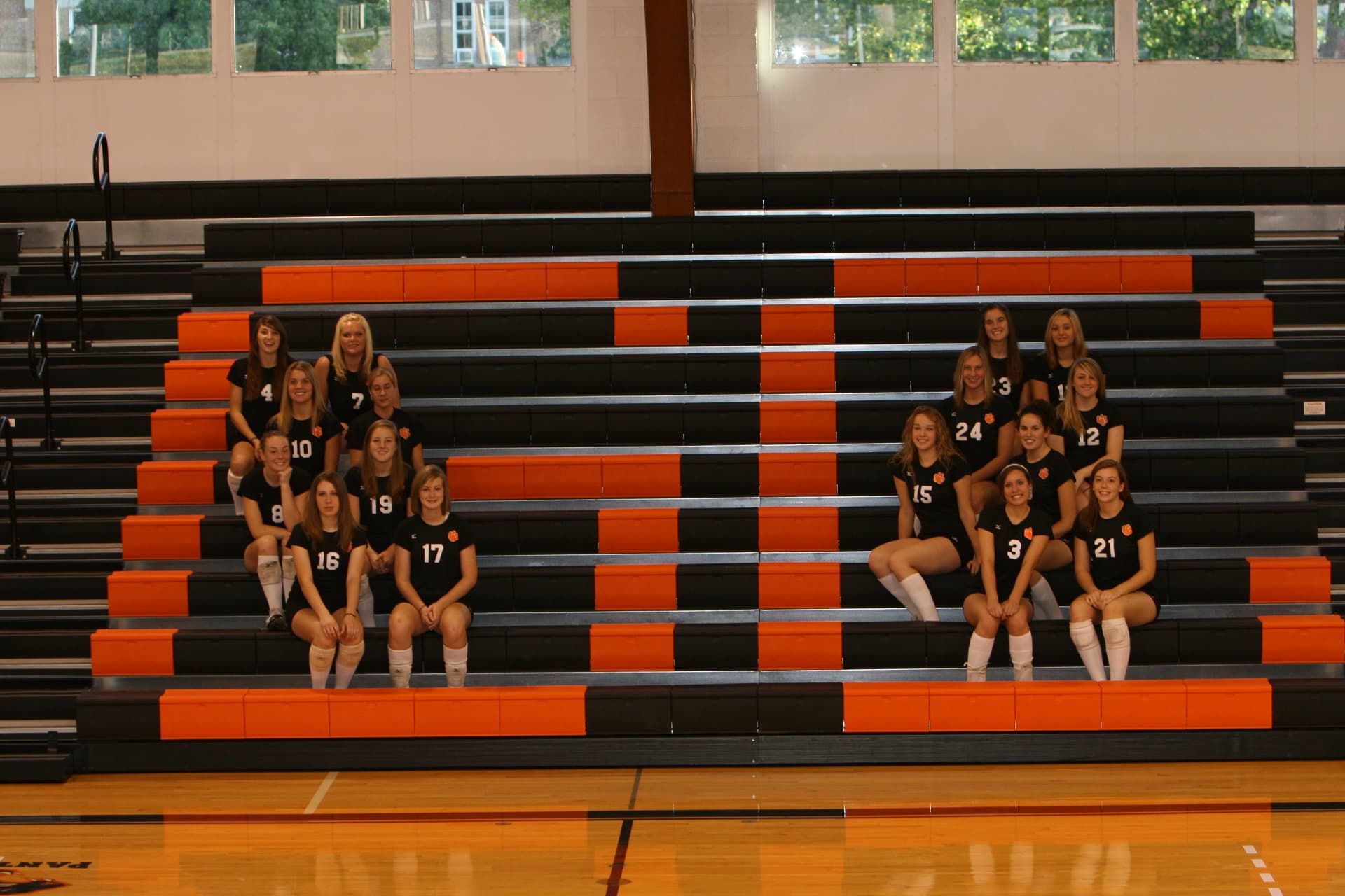 Greenville volleyball team seated across the orange and black bleachers of the gymnasium.